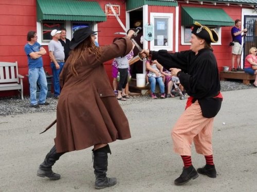 Two performers in pirate costumes engage in a playful sword fight on a street, with an audience watching nearby.