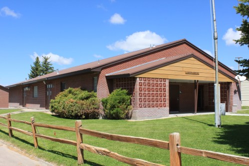 A brick building with a wooden entrance, surrounded by green grass and a wooden fence, under a blue sky with clouds.