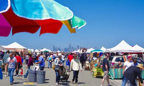 A bustling outdoor market with colorful umbrellas, tents, and people shopping, with a city skyline in the background.