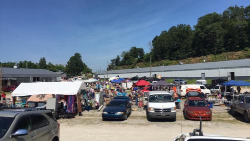 A bustling outdoor market with numerous tents, cars, and people browsing among various stalls on a sunny day.