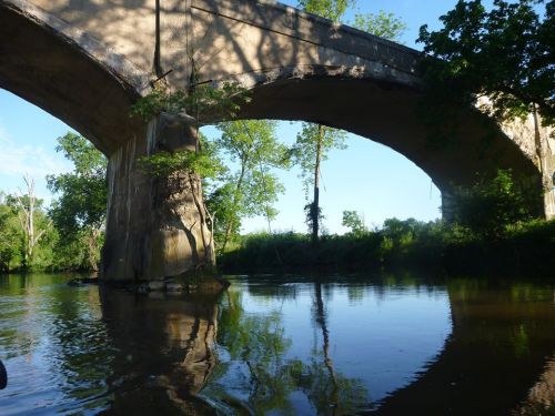 A serene river scene featuring two large, weathered bridges reflected in the calm water, surrounded by lush greenery.