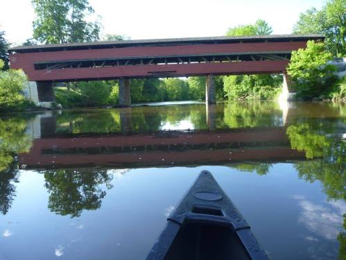 A canoe view of a red covered bridge reflecting in calm water, surrounded by lush green trees.