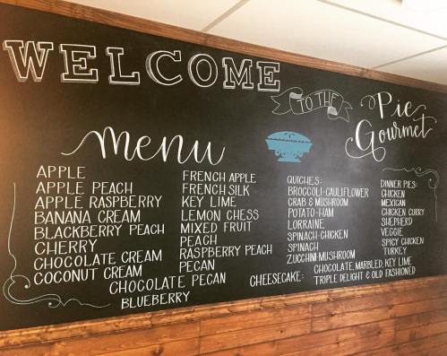 Chalkboard menu at Pie Gourmet listing various pies, quiches, and cheesecakes with flavors like apple, blueberry, and chocolate.