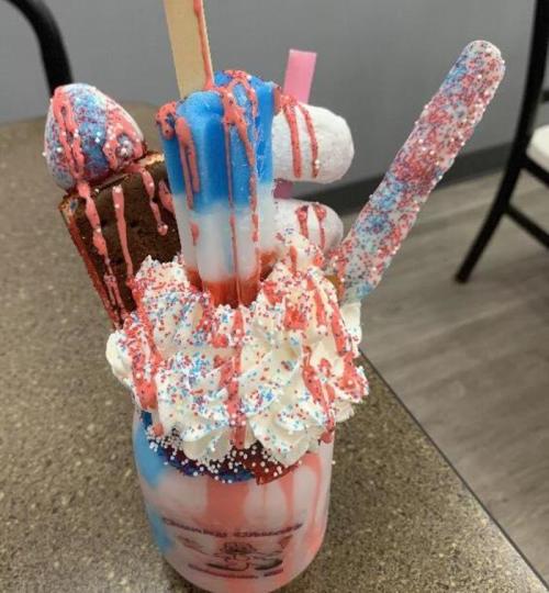 A colorful milkshake topped with whipped cream, sprinkles, and various sweets, including marshmallows and a cookie.