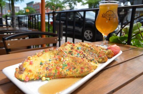 A plate of colorful, cereal-crusted French toast with syrup and a strawberry, alongside a glass of beer on a table.