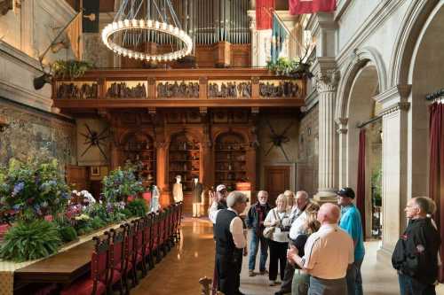 A group of people gathers in a grand hall with a long dining table, ornate decor, and a large chandelier overhead.