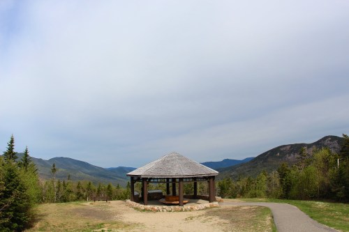 A gazebo overlooks a scenic mountain landscape under a cloudy sky, with a winding path leading towards it.