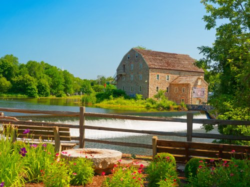 A scenic view of a stone mill by a river, surrounded by greenery and colorful flowers under a clear blue sky.