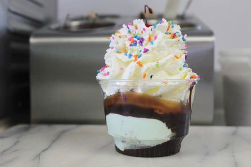 A cup of mint chocolate chip ice cream topped with whipped cream and colorful sprinkles, sitting on a marble surface.