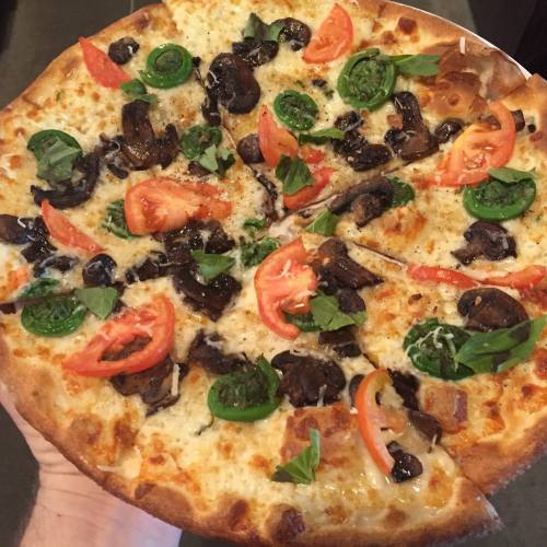 A hand holding a pizza topped with mushrooms, tomatoes, basil, and green herbs on a golden crust.