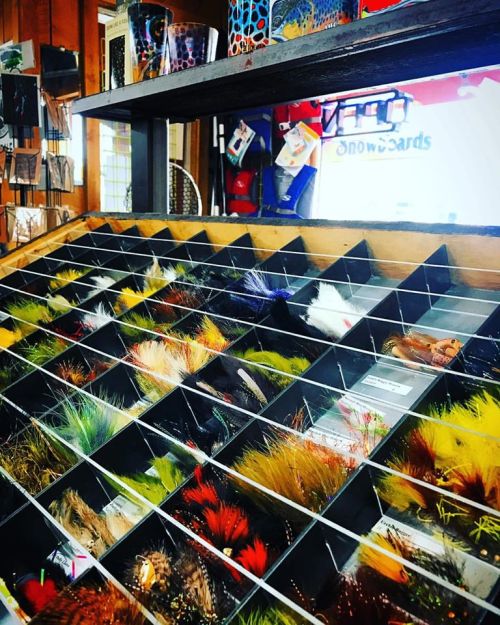 A display of colorful fishing flies organized in a wooden rack, with various patterns and textures visible.