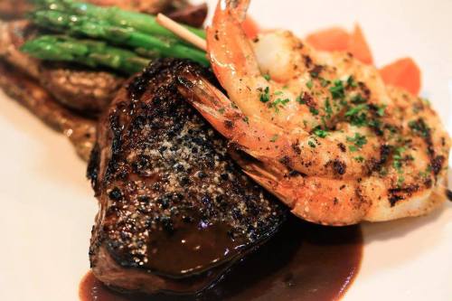 A plated dish featuring grilled shrimp, a juicy steak, asparagus, and garnished with herbs.