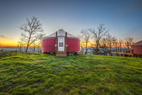 A red yurt on a grassy hill at sunset, surrounded by bare trees and a scenic view of the water.