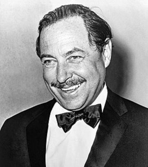 A smiling man in a tuxedo with a bow tie, featuring a mustache and slicked-back hair, against a plain background.