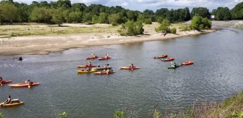 A group of people kayaking on a calm river surrounded by greenery and a sandy shore under a partly cloudy sky.