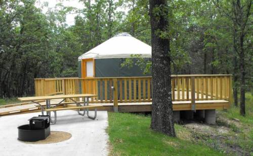A wooden deck surrounds a green yurt in a forested area, with a picnic table and fire pit nearby.