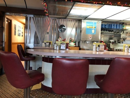 A cozy diner interior featuring a circular counter with red stools, festive decorations, and a kitchen area in the background.