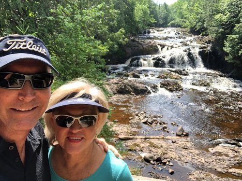 A man and woman pose for a selfie in front of a scenic waterfall surrounded by lush greenery.
