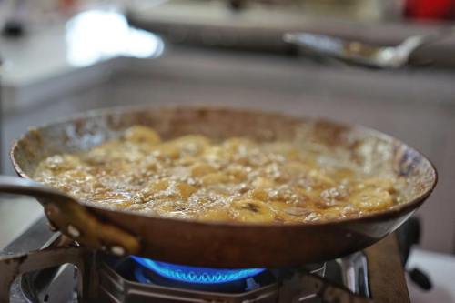 A frying pan with sliced bananas cooking in hot oil on a stovetop, with a blue flame visible underneath.