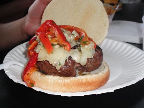 A close-up of a juicy burger topped with melted cheese and colorful roasted peppers, served on a paper plate.