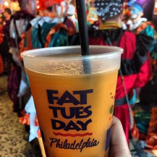 A hand holds a drink with "Fat Tuesday Philadelphia" printed on it, with colorful costumed figures blurred in the background.