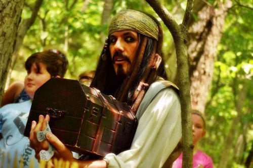 A person dressed as a pirate holds a treasure chest, surrounded by children in a forest setting.