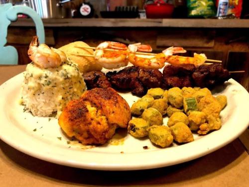 A plate of assorted seafood and meats, including shrimp, rice, and fried okra, served with cornbread and skewers.