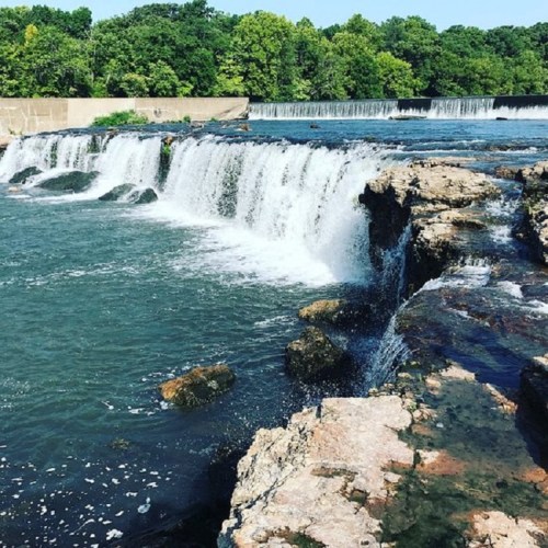 A scenic view of a waterfall cascading over rocky ledges, surrounded by lush green trees and a clear blue sky.