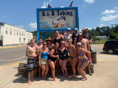A group of people in swimwear poses in front of a sign for S & B Tubing on a sunny day.