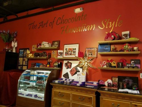 A vibrant shop interior featuring chocolate displays, shelves of treats, and a decorative wall with "The Art of Chocolate Hawaiian Style."