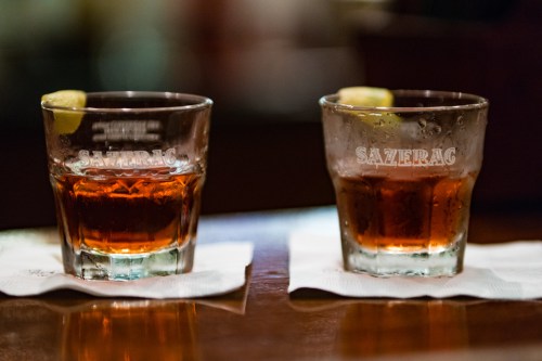 Two Sazerac cocktails in short glasses, garnished with lemon, on white napkins against a blurred background.
