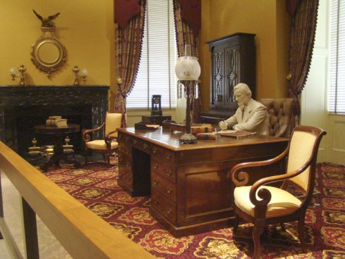A historic office setting with a wooden desk, chairs, and a statue of a man writing, surrounded by elegant decor.