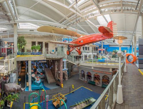 A vibrant indoor museum space featuring a vintage airplane suspended from the ceiling, with interactive exhibits below.