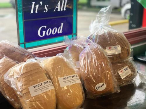 Loaves of white and brown bread in plastic bags displayed on a counter, with a sign reading "It's All Good" in the background.