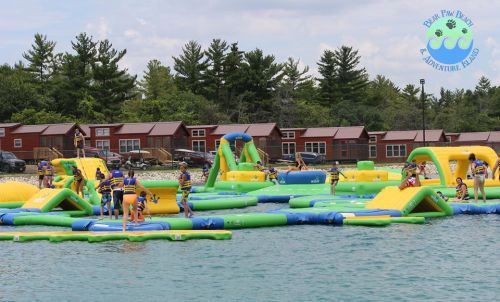 A colorful inflatable water park with people playing and enjoying activities on a lake, surrounded by trees and cabins.