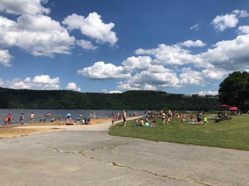 A busy beach scene with people swimming and relaxing on the shore under a blue sky with fluffy clouds.