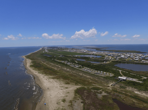 Aerial view of a coastal area with sandy shores, water, and a small town in the distance under a clear blue sky.