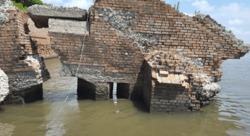 Ruined brick structure partially submerged in water, showing erosion and decay, with greenery in the background.