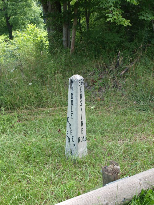 A weathered stone mile marker indicating "Middle Creek 30" and "Erskine Road 324" in a grassy area.