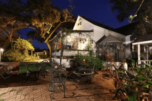 A cozy backyard at night, featuring a patio with metal furniture, lush greenery, and a well-lit house in the background.