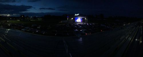A drive-in movie screen illuminated at night, with cars parked in front and a dark sky overhead.