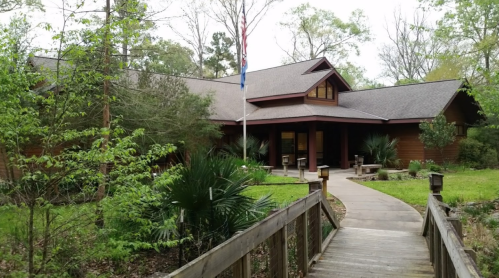 A wooden building surrounded by greenery, with a pathway leading to the entrance and flags in front.