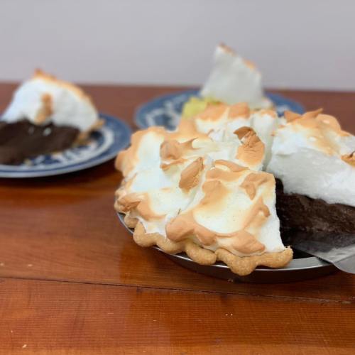 A slice of chocolate meringue pie on a plate, with a whole pie in the background, both on a wooden table.