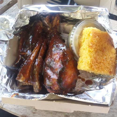 A takeout box containing two barbecued chicken legs and a piece of cornbread, all wrapped in foil.