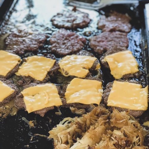 Grilled burgers with melted cheese on a hot griddle, alongside sautéed onions.