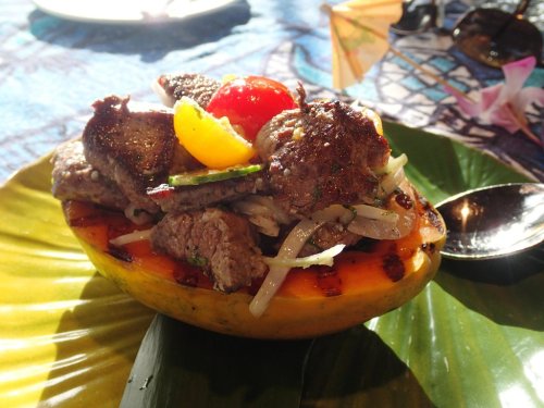 A colorful dish of grilled beef served in a halved papaya, garnished with vegetables and a cherry tomato.