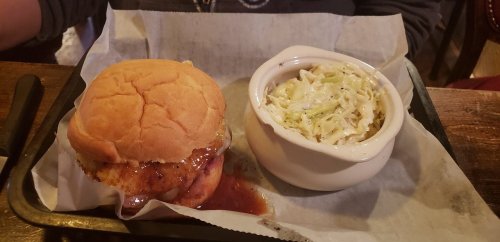 A BBQ chicken sandwich with sauce on a bun, served with a side of coleslaw in a small bowl on a tray.