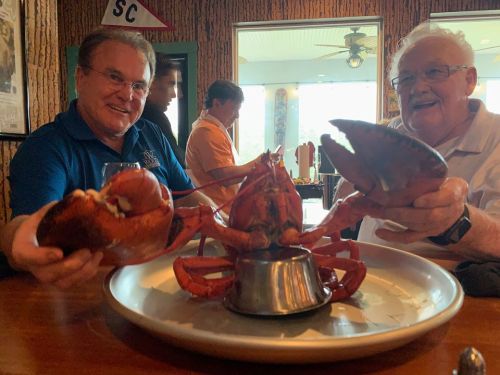 Two men at a restaurant hold up lobsters over a large plate, smiling and enjoying their meal together.