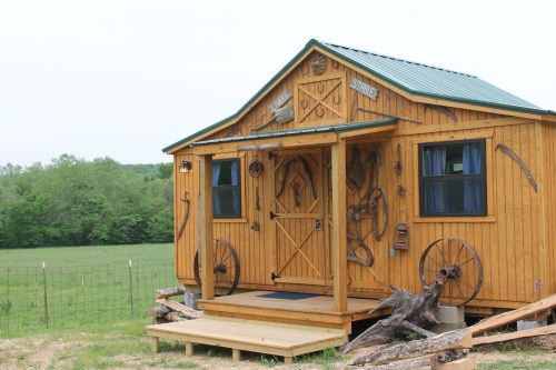 A rustic wooden cabin with a green metal roof, featuring decorative tools and a porch, set in a grassy landscape.