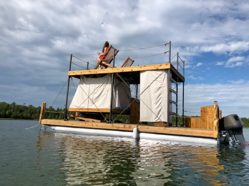 A floating structure on water with a person relaxing in a chair, surrounded by trees and a cloudy sky.